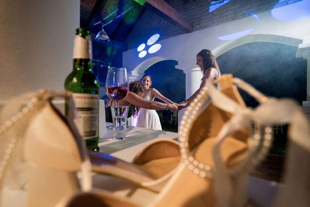 Two women celebrate at a bar with red wine glasses, pearl heels in foreground, green wine bottle nearby.
