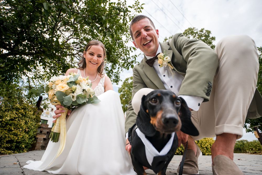 Bride and groom pose with their dachshund dog dressed in a tuxedo on their wedding day.