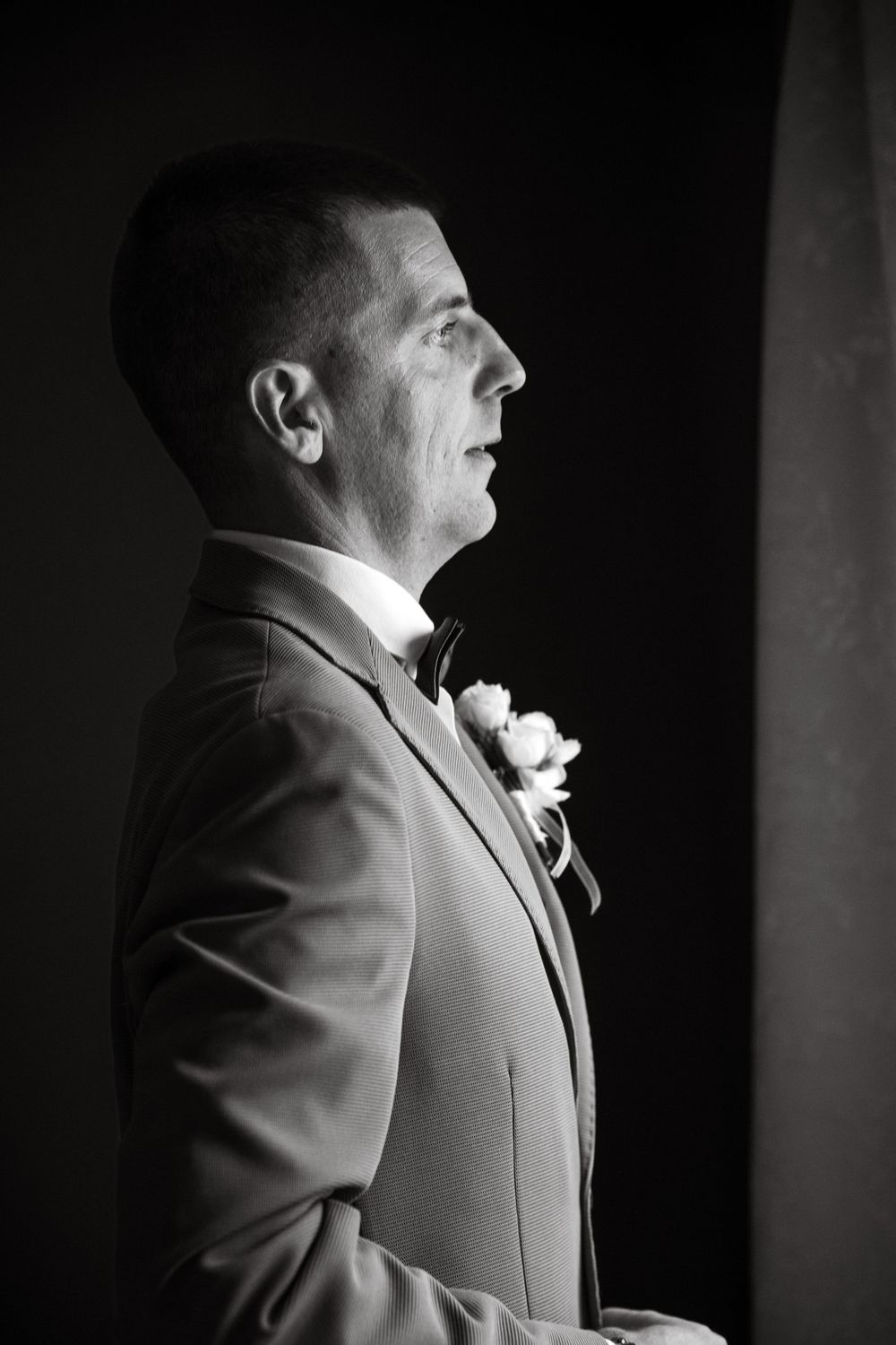 Black and white portrait of a groom in a suit with boutonniere, gazing upward in dramatic side lighting.