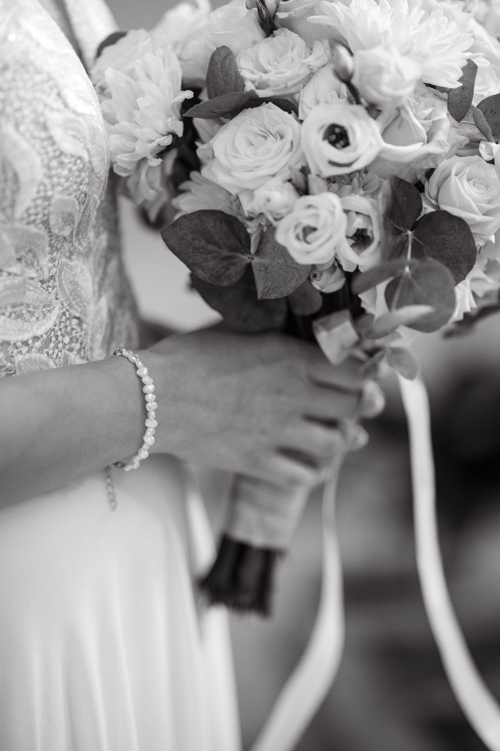 Black and white photo of a bride holding a wedding bouquet of roses and eucalyptus, wearing a pearl bracelet.