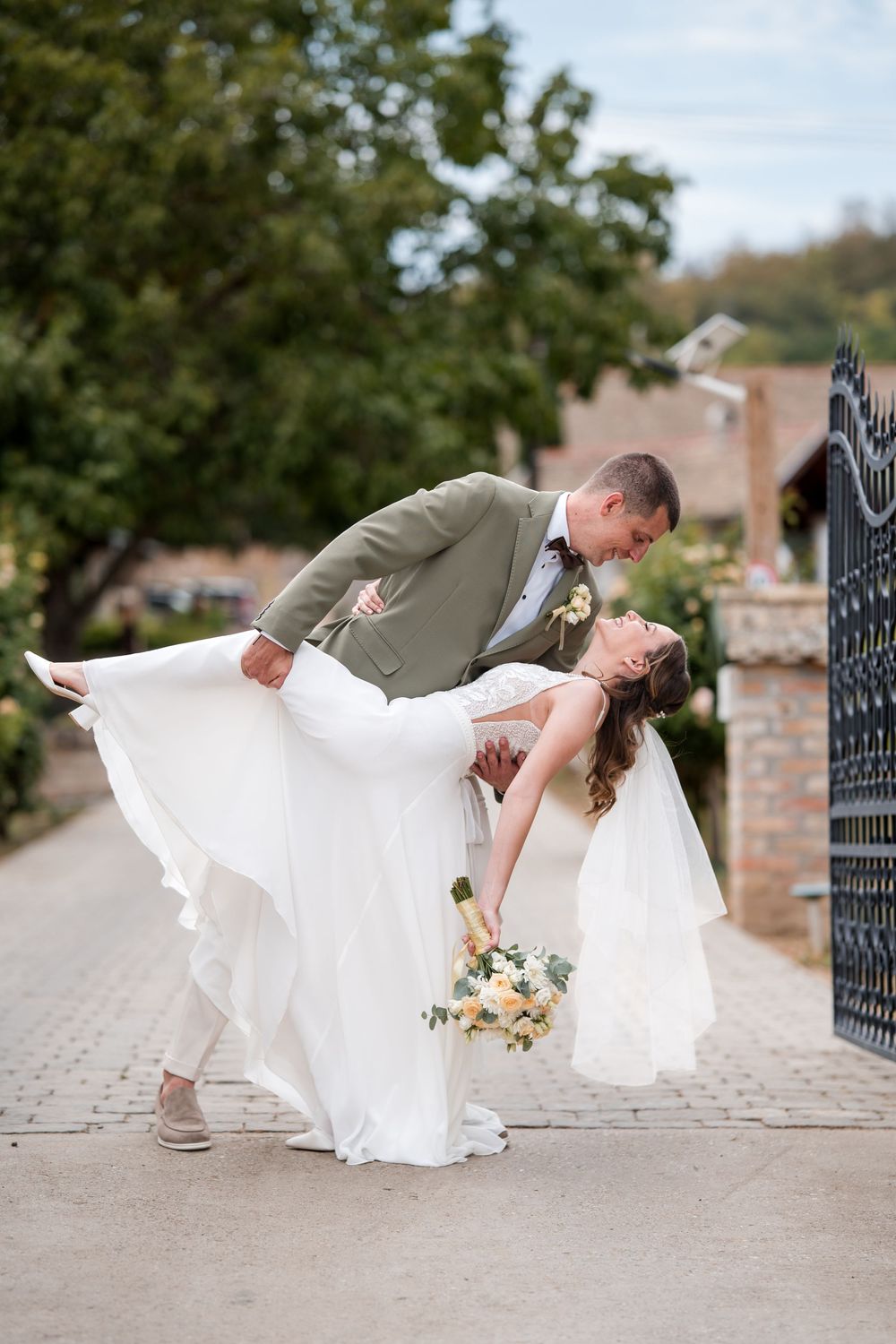 Groom in olive suit dips bride in white gown for a romantic kiss near ornate iron gate at outdoor wedding.