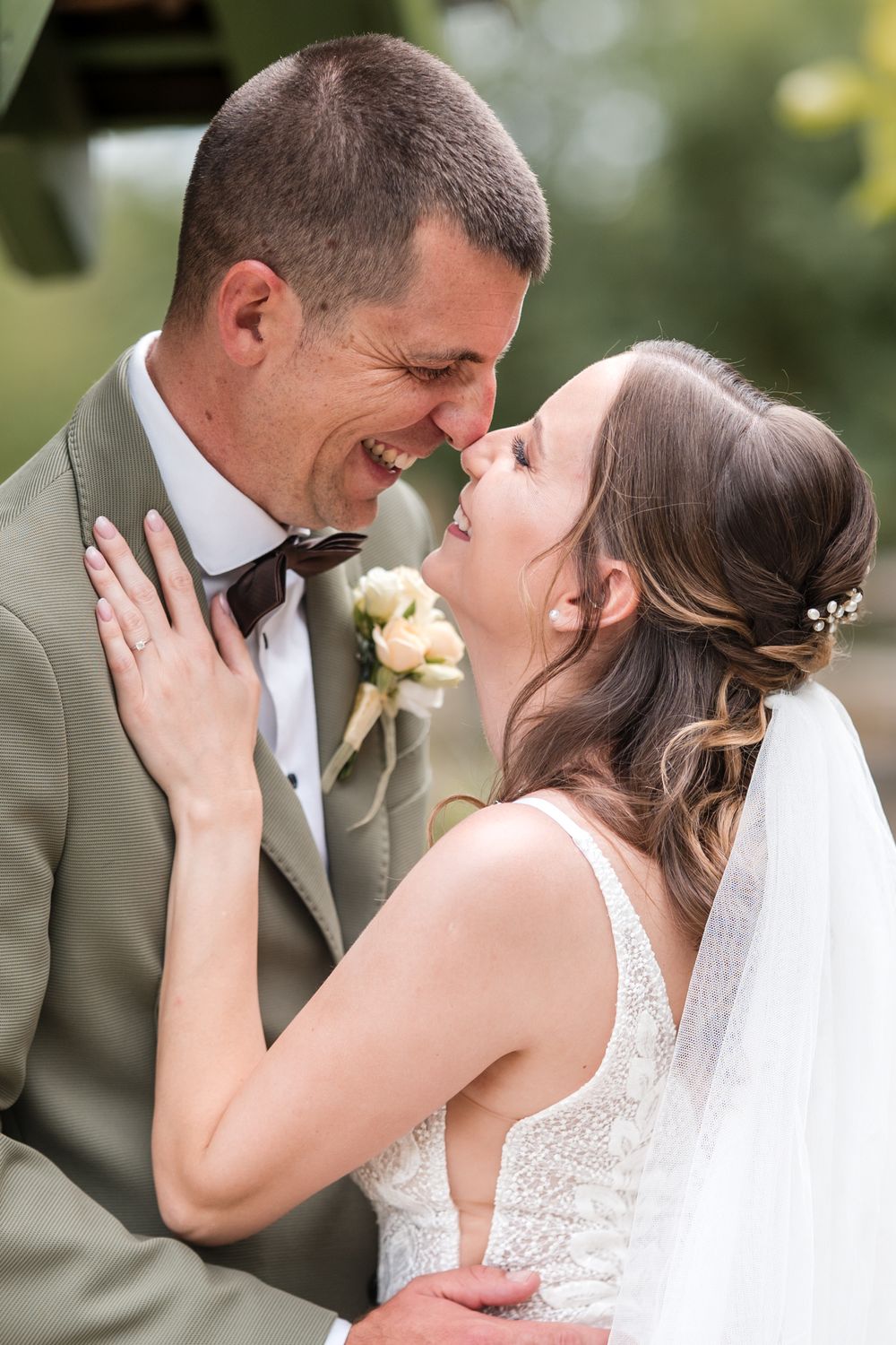 Smiling bride and groom touching foreheads in an intimate wedding moment, groom in sage suit, bride in lace gown with veil.