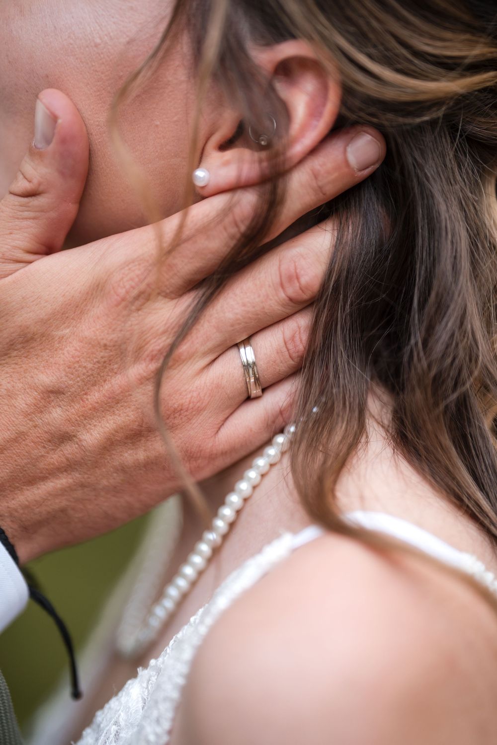 Groom with wedding ring tenderly holding brides neck during romantic wedding kiss, pearl necklace visible.