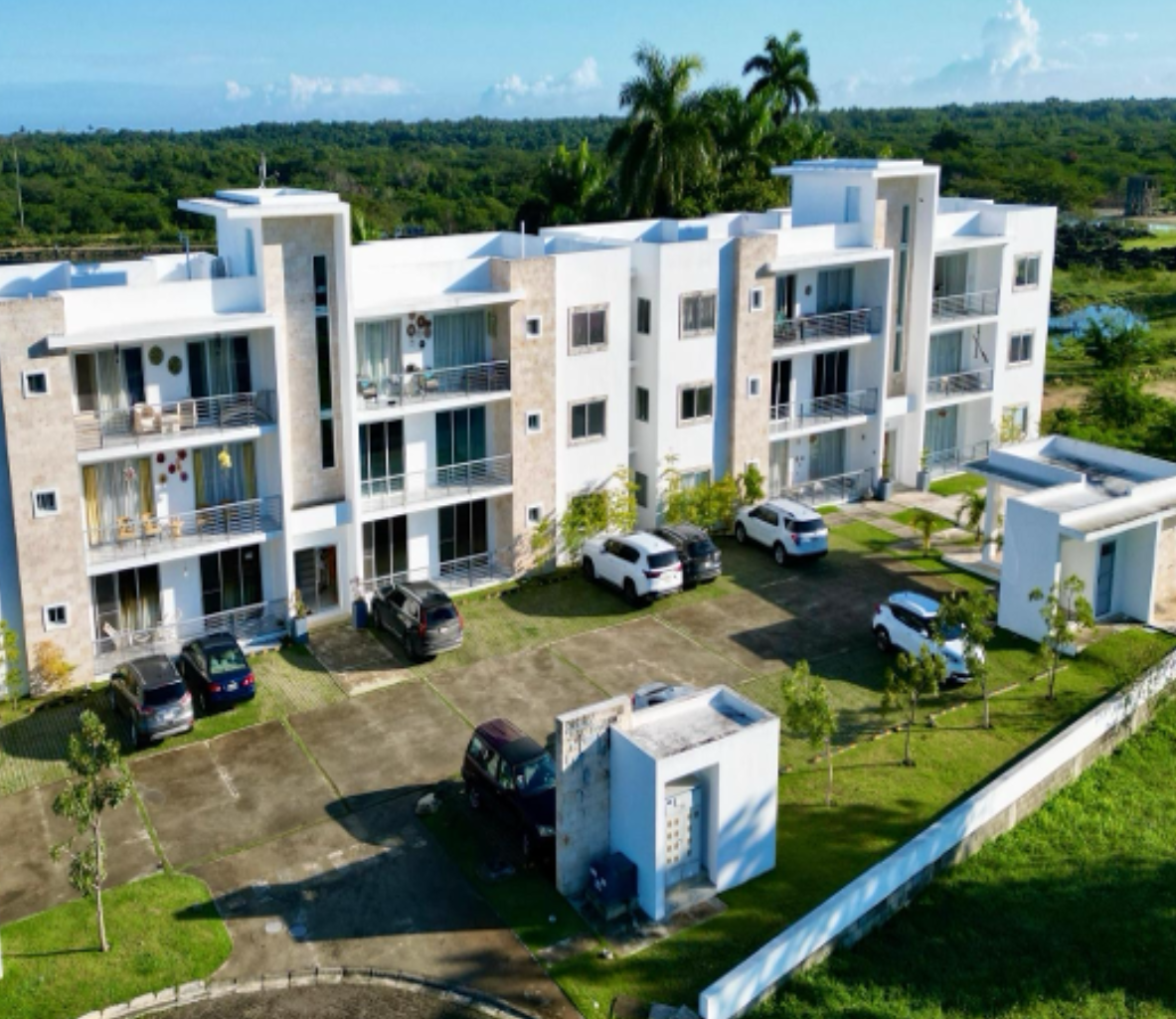 Aerial view of white three-story apartment complex with green lawn, parking spaces, and tropical surroundings.