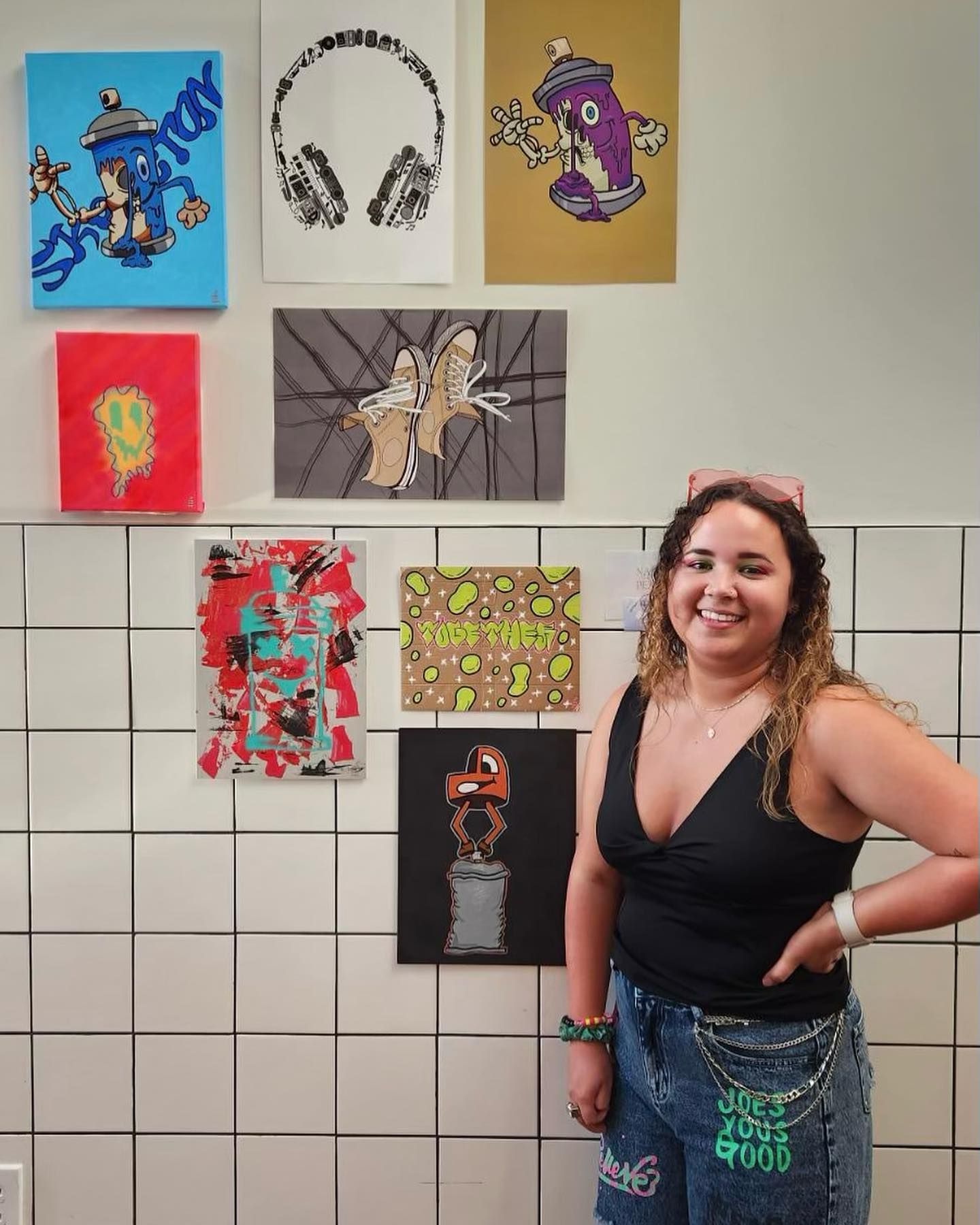 Woman smiling beside colorful graffiti-style artwork displayed on white tiled wall at an art show.