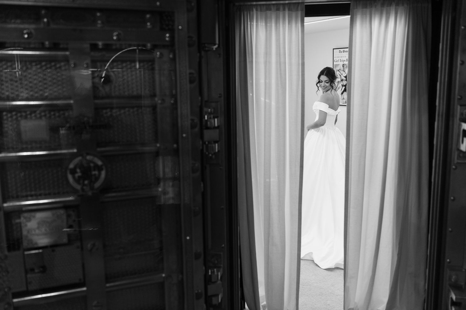 Bride in white wedding gown standing in doorway beside vintage bank vault door, black and white photo.
