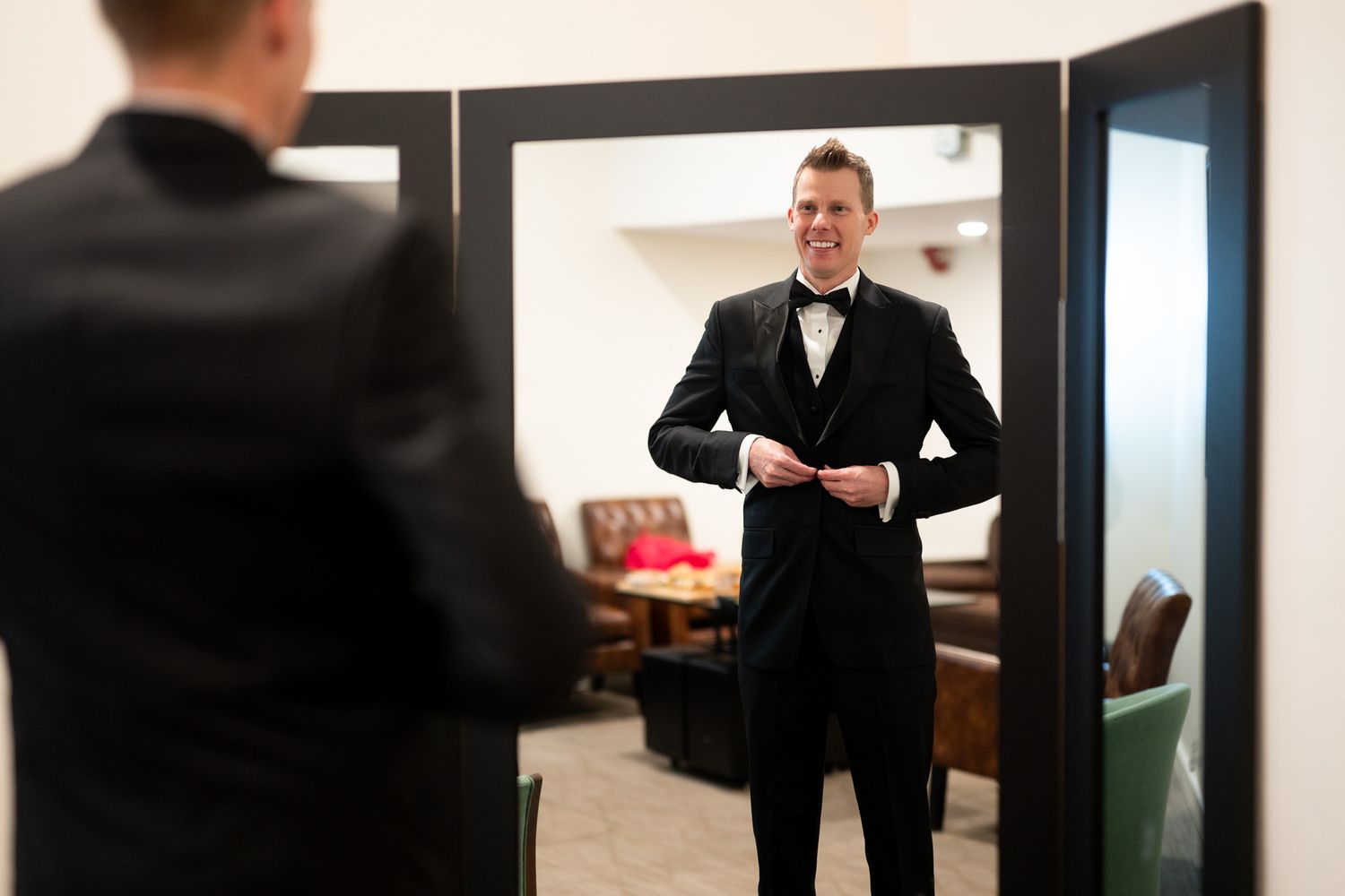 Groom in black tuxedo and bow tie buttoning jacket while smiling at his reflection in a mirror.