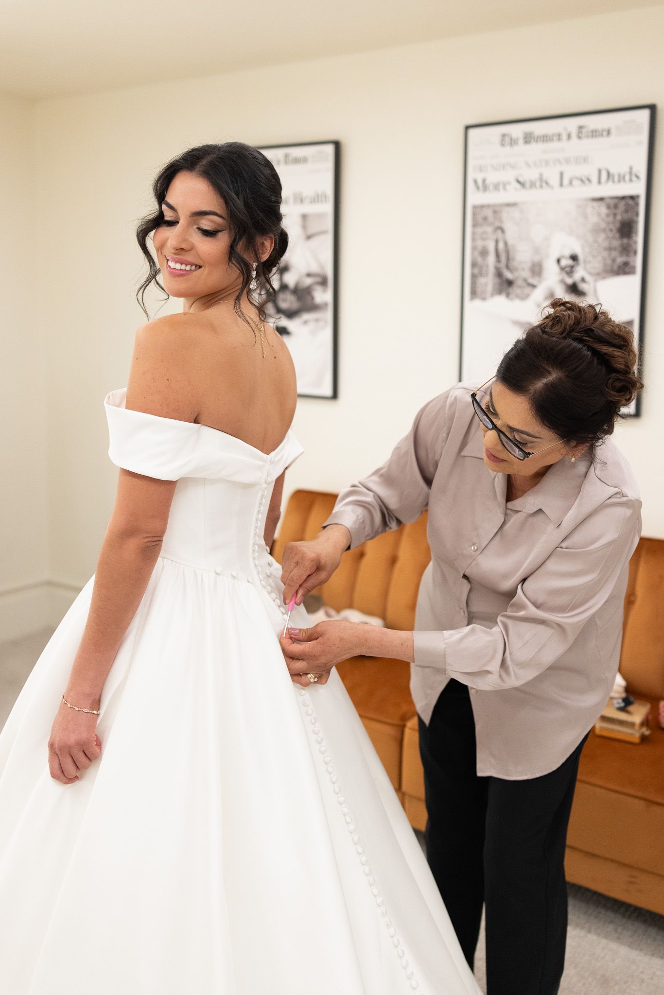Bride in off-shoulder white ball gown smiles as woman helps lace up the back of her wedding dress.