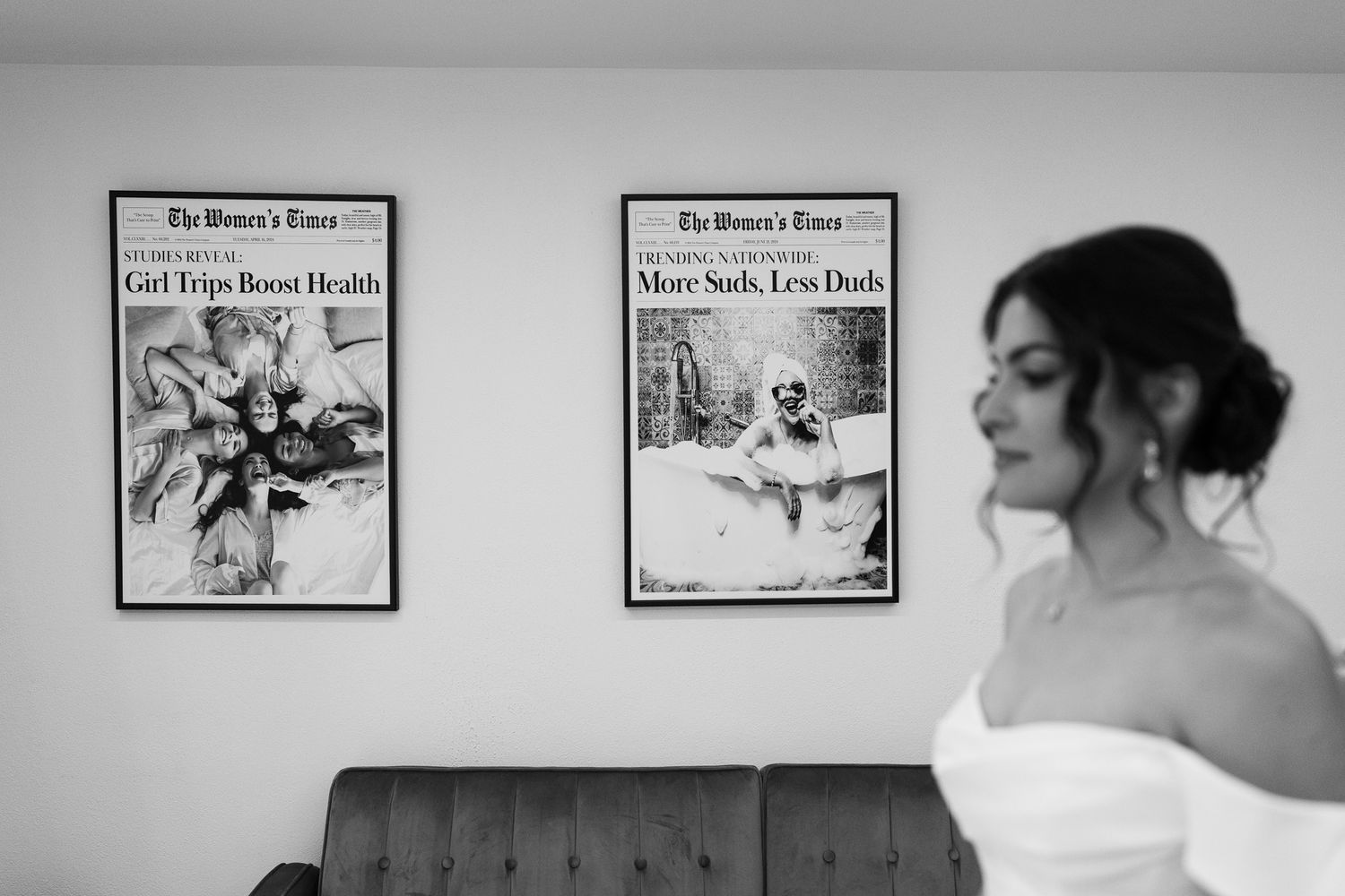 Black and white photo of a bride in a strapless gown standing near framed newspaper prints on a wall.