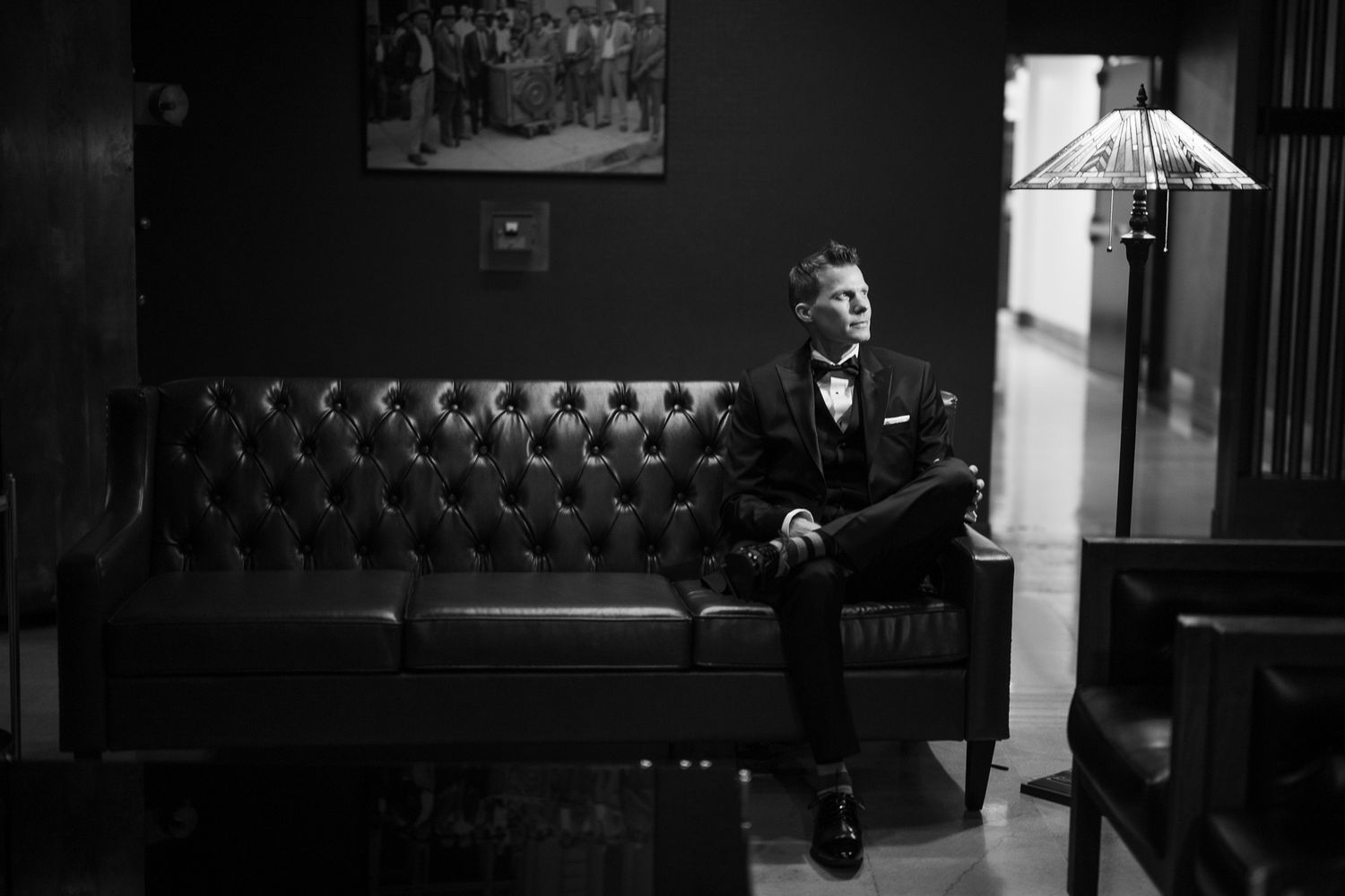 A dapper man in a black tuxedo sits pensively on a tufted leather sofa in an elegant dimly lit room.