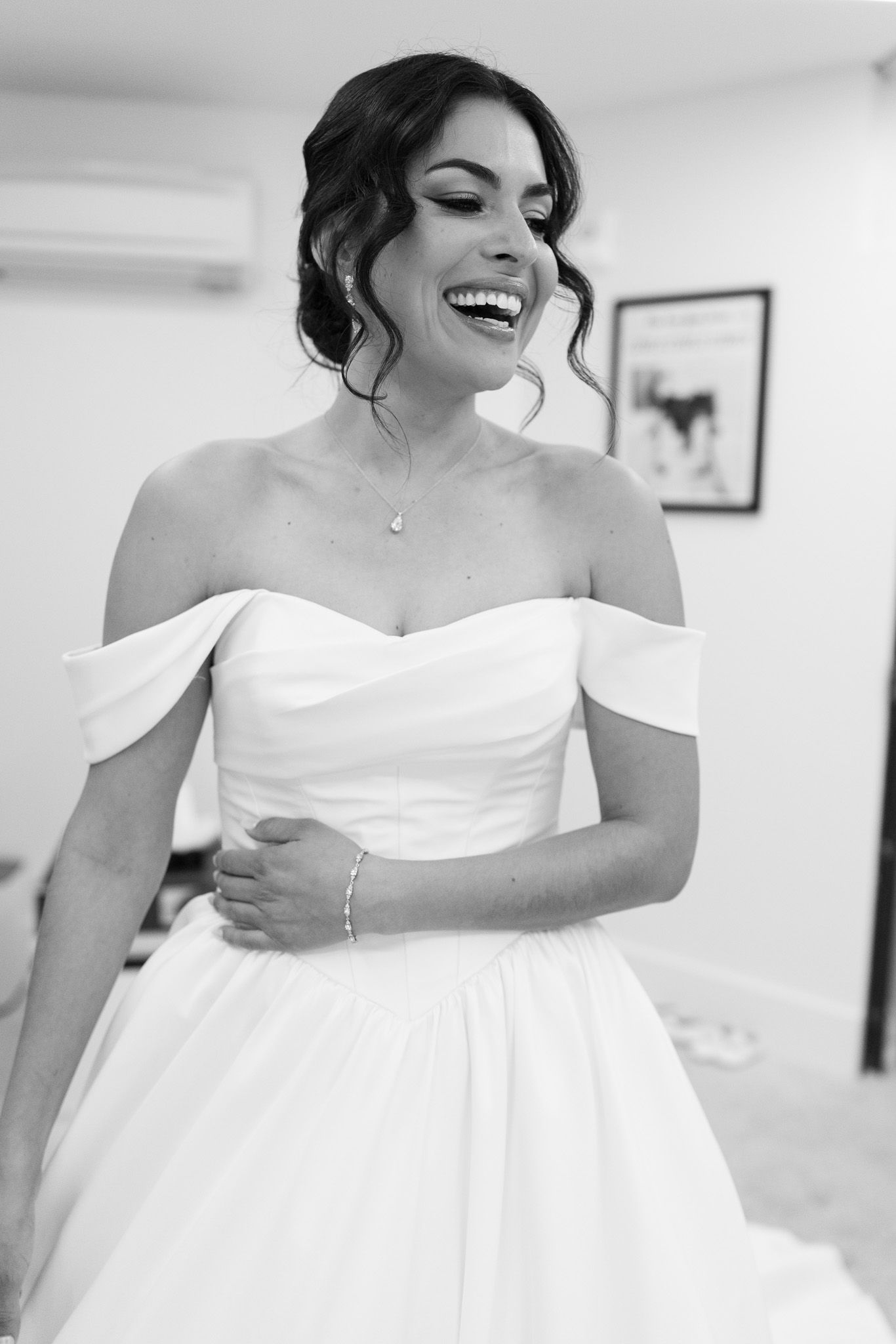 Black and white photo of a laughing bride in an off-shoulder wedding gown with delicate necklace and bracelet.