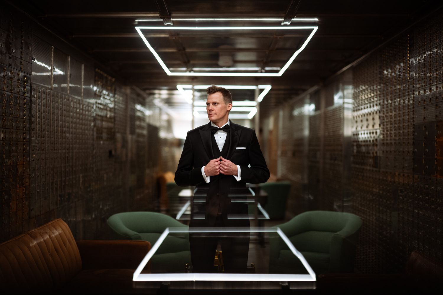 Elegant man in tuxedo and bow tie stands at a glass table under geometric neon lights in a stylish interior.