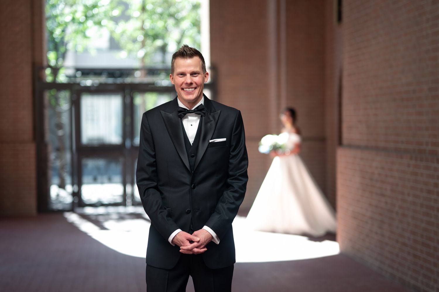 Smiling groom in black tuxedo stands outdoors with bride in white wedding dress visible in background.