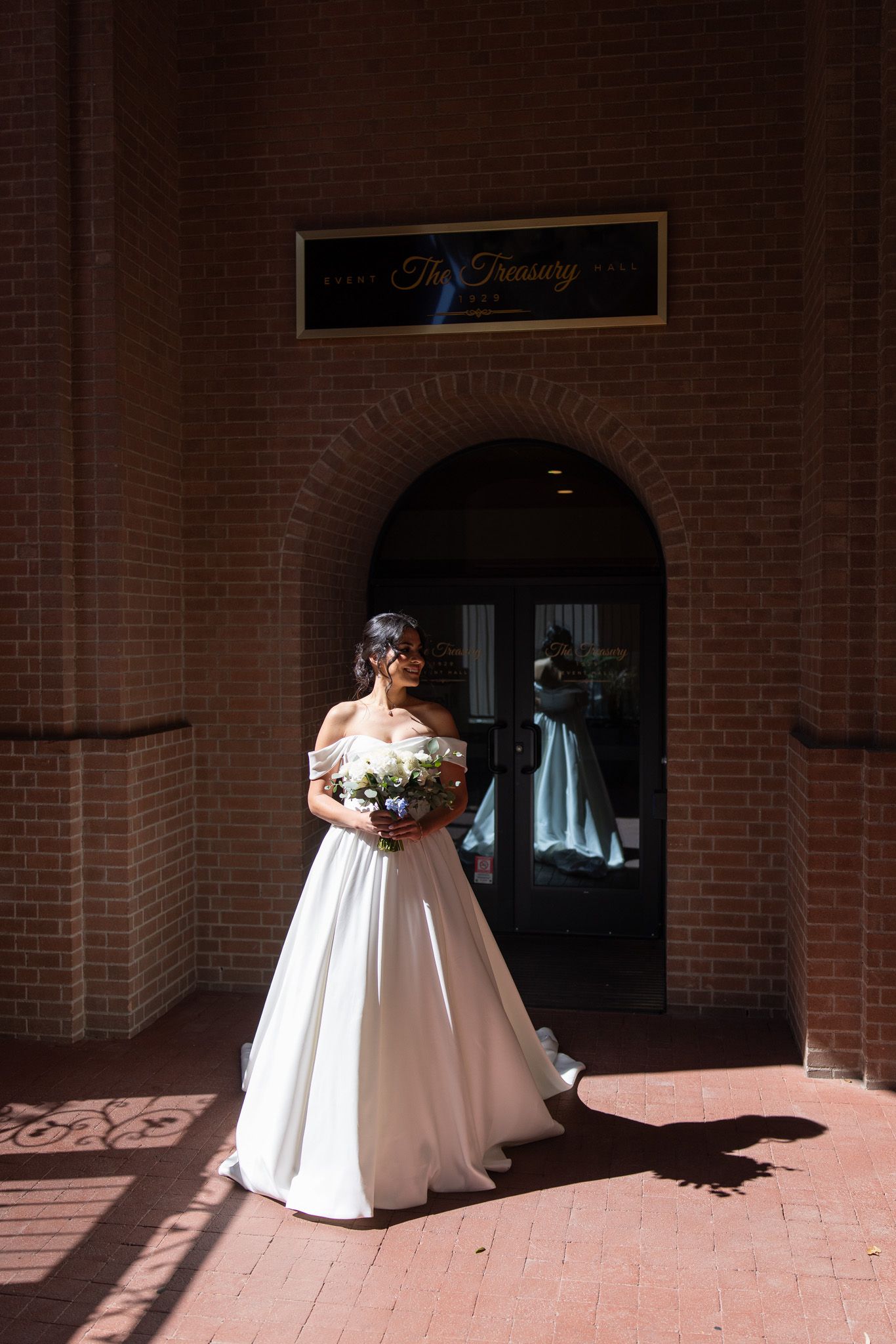 Bride in off-shoulder white ball gown holding bouquet outside The Treasury brick building entrance.