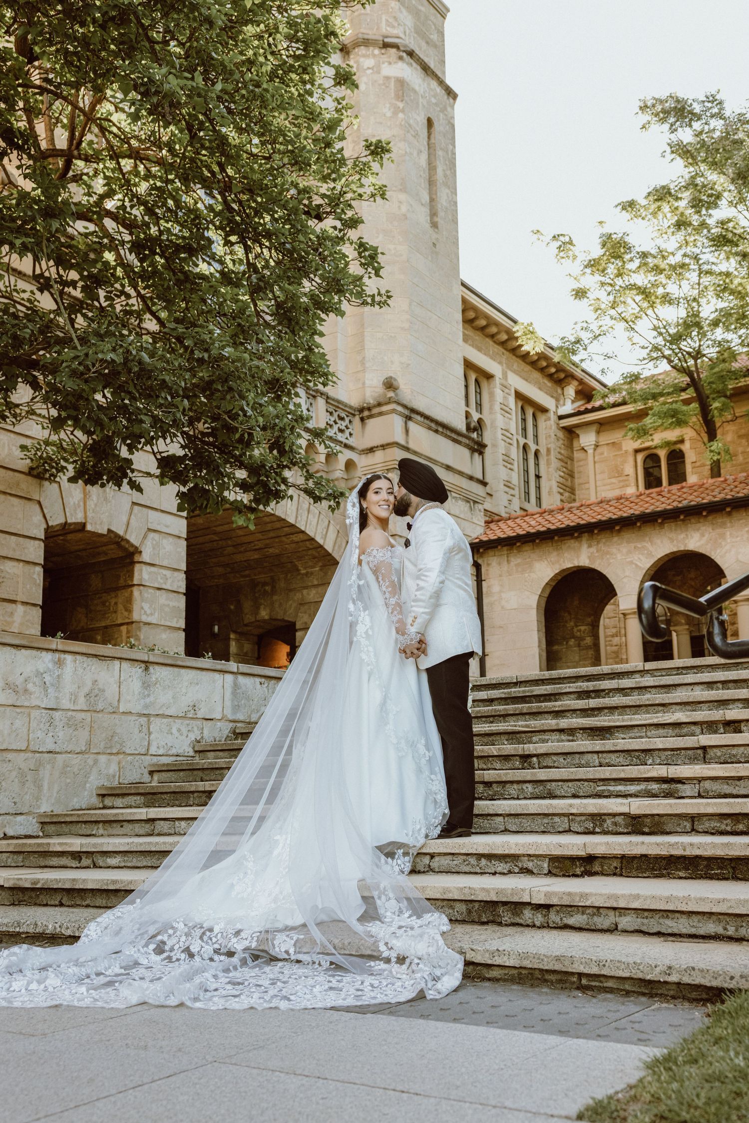 Bride in lace gown with cathedral veil and groom in white tuxedo share a kiss on stone steps of elegant venue.