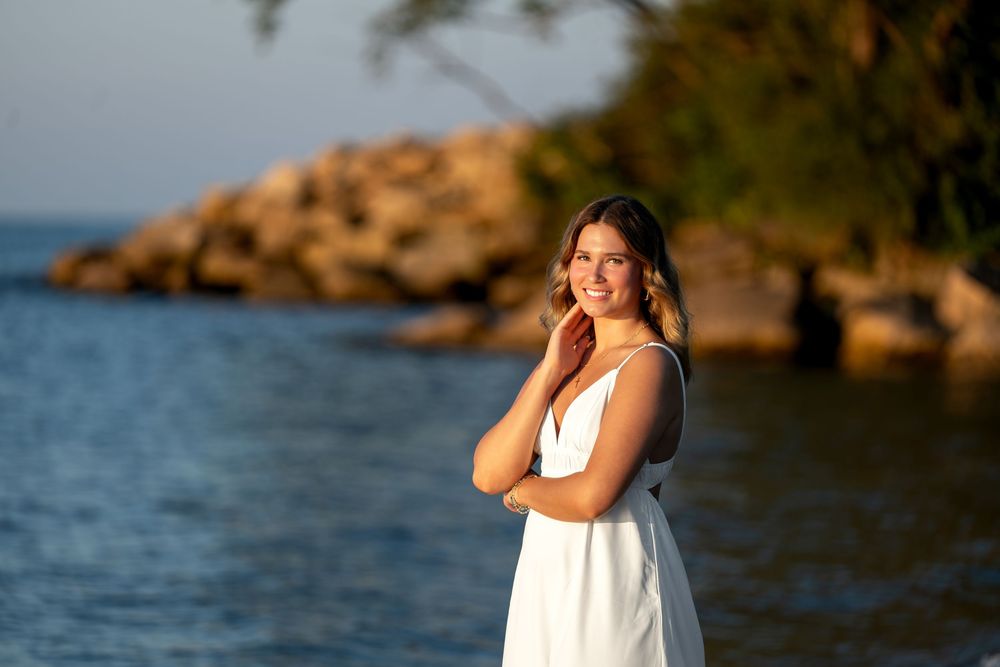 Young woman in white dress smiling by the water at sunset with rocky shore and trees in background.