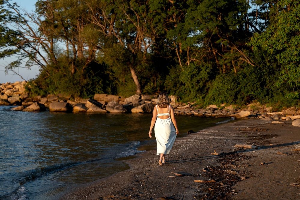 Woman in white outfit walking along a rocky tropical beach at golden hour with lush green trees in background.