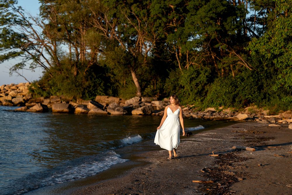 Woman in white dress walking along a rocky tropical beach shore at golden hour sunset.