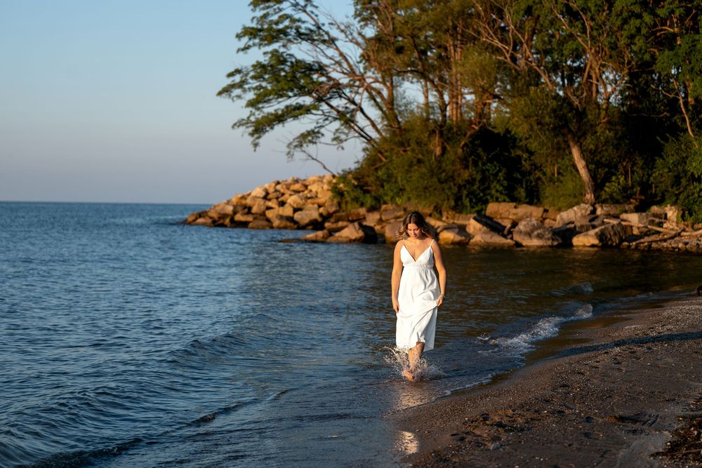 Woman in white dress wading along a serene rocky shoreline at golden hour with lush green trees in the background.