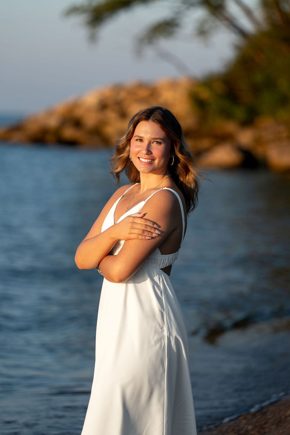 Smiling young woman in white dress standing on rocky beach at golden hour sunset.