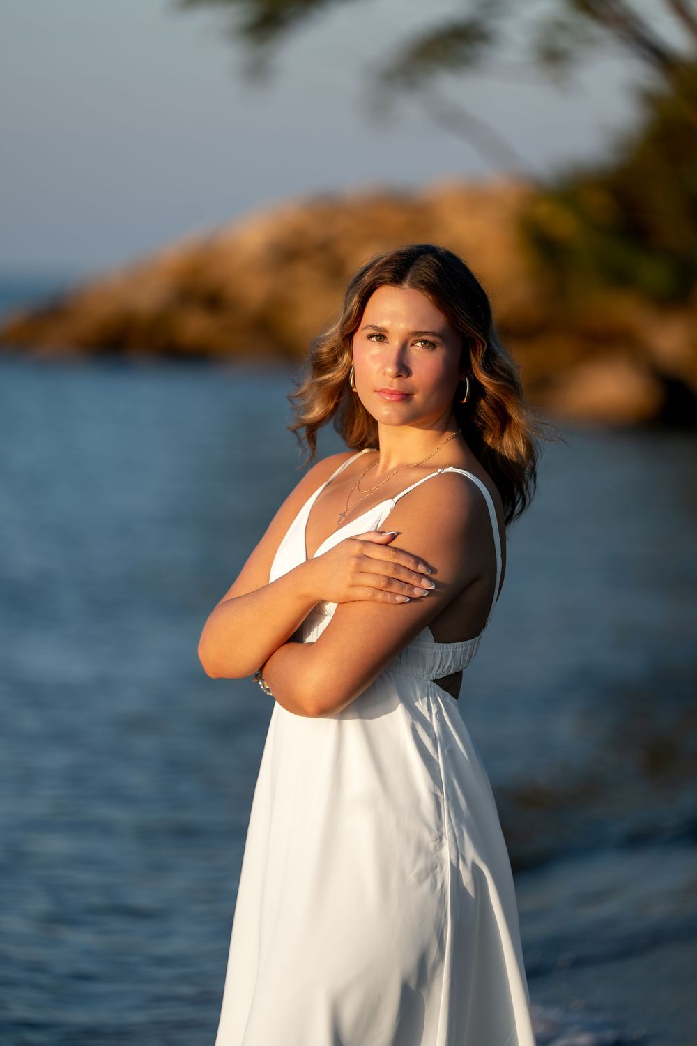 Young woman in white dress with arms crossed poses by rocky seaside at golden hour sunset.
