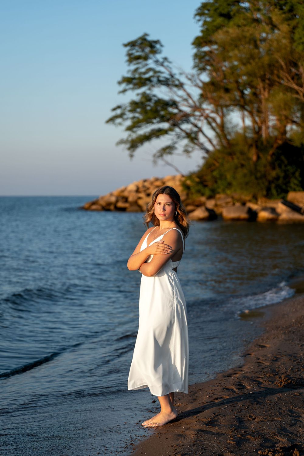Barefoot woman in white maxi dress stands on sandy beach shore at sunset with trees behind.