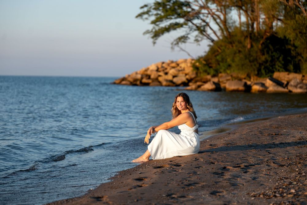 Woman in white dress sitting on sandy beach shore at golden hour with rocky coastline and trees in background.