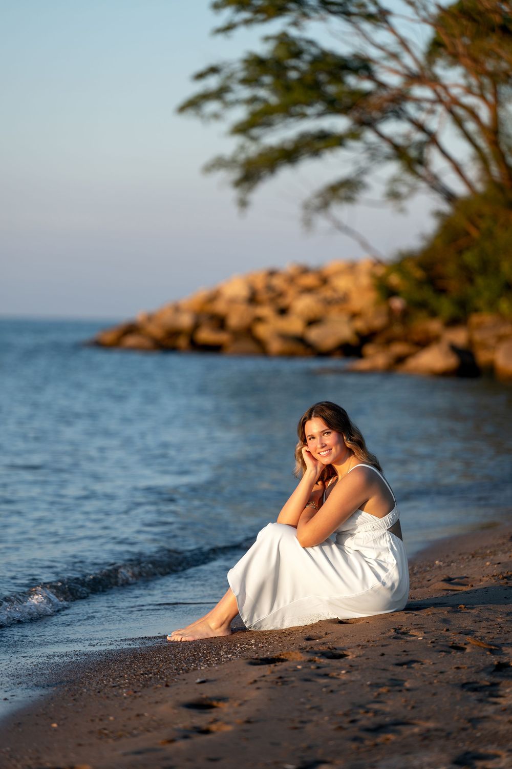 Smiling woman in white dress sitting on sandy beach at sunset with rocky shoreline and trees in background.