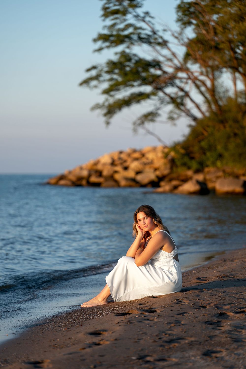 Young woman in white dress sitting barefoot on sandy beach at golden hour with rocky shoreline and trees.