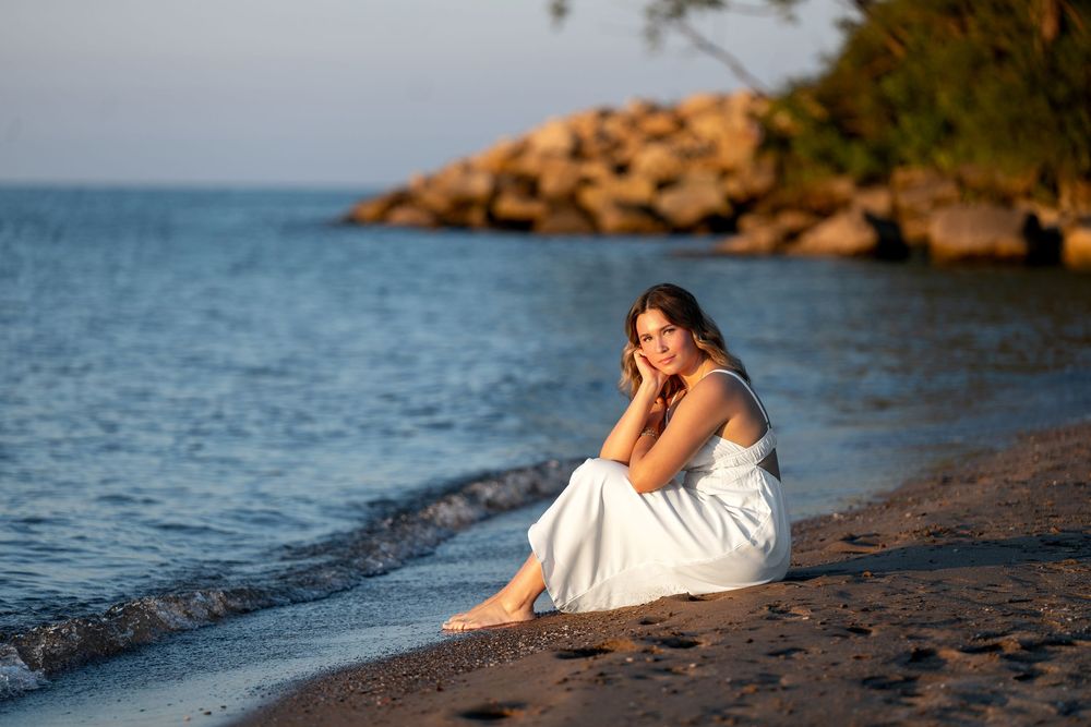 A young woman in a white dress sits barefoot on a sandy beach at golden hour, gazing thoughtfully toward the sea.