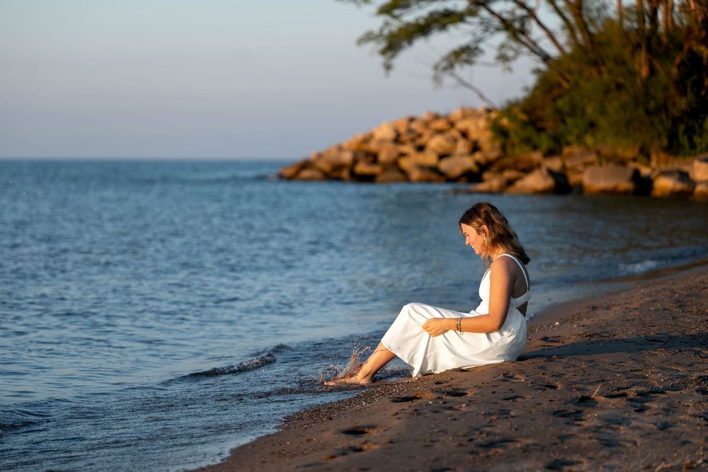 Woman in white dress sitting on sandy beach shore at sunset near rocky coastline and calm blue water.