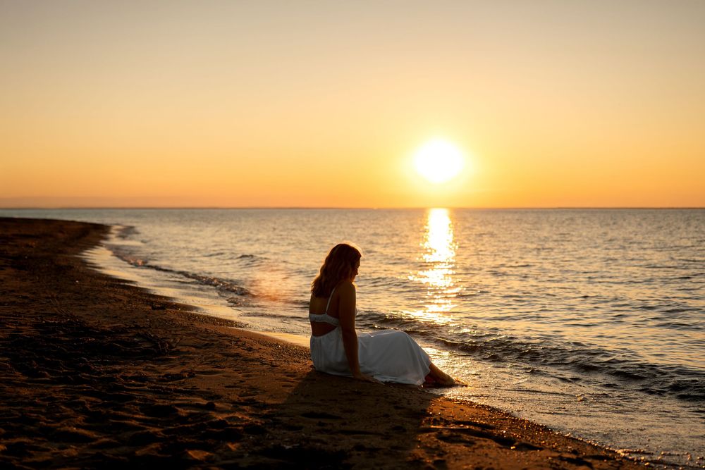 Woman in white dress sitting alone on sandy beach during stunning golden sunset.