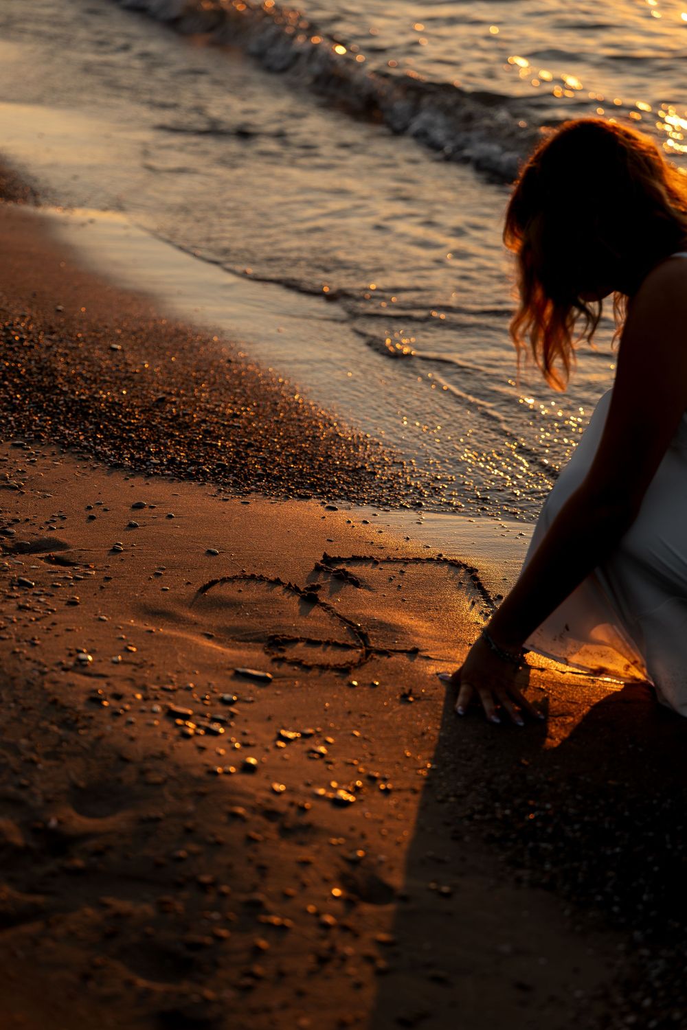 Woman in white dress drawing a heart in wet sand on a beach at golden sunset.
