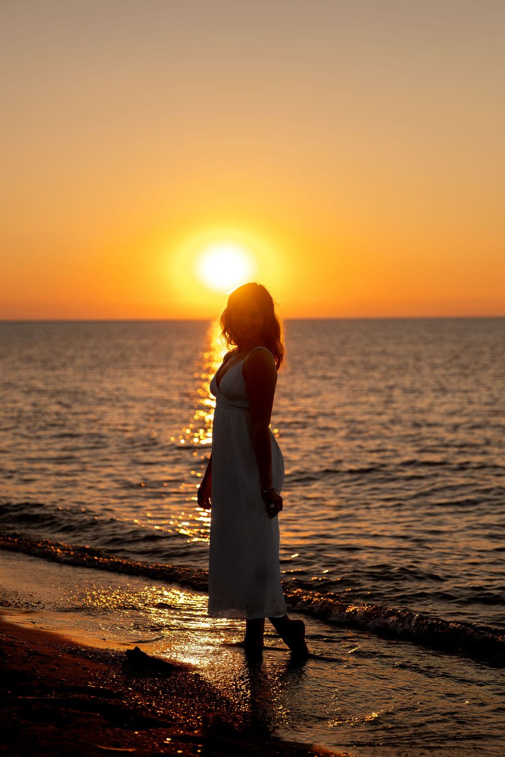 Elegant silhouette of woman in white dress standing on beach shoreline against vibrant golden sunset sky.