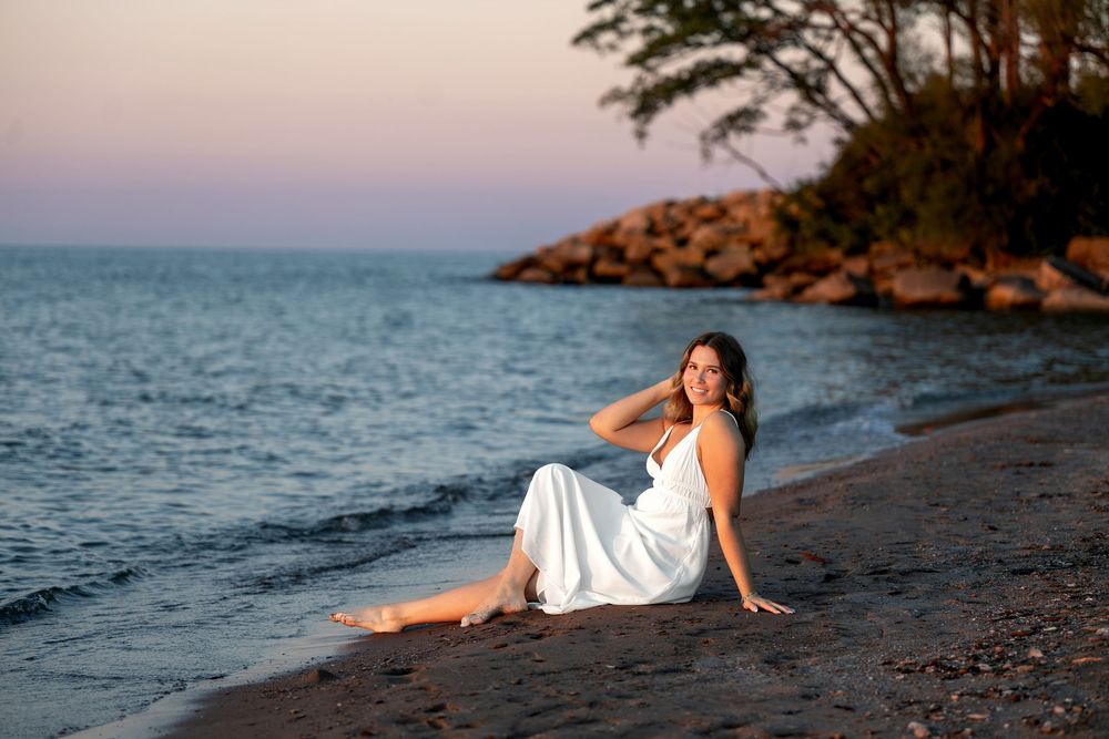 Woman in white dress sitting on sandy beach at pink sunset near rocky shore.
