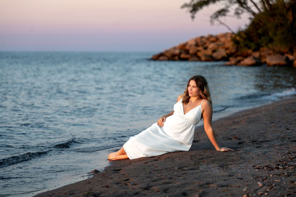 Woman in white dress sitting gracefully on beach at dusk with calm sea and rocky shore behind her.