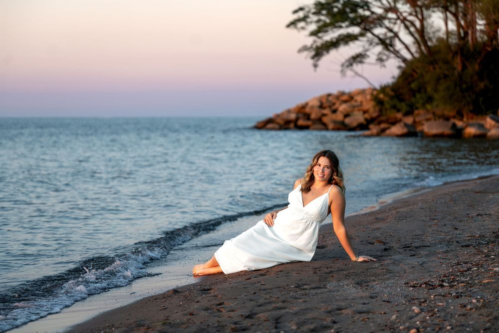 Woman in white dress sitting on sandy beach at dusk, relaxing by calm ocean with rocky shore backdrop.