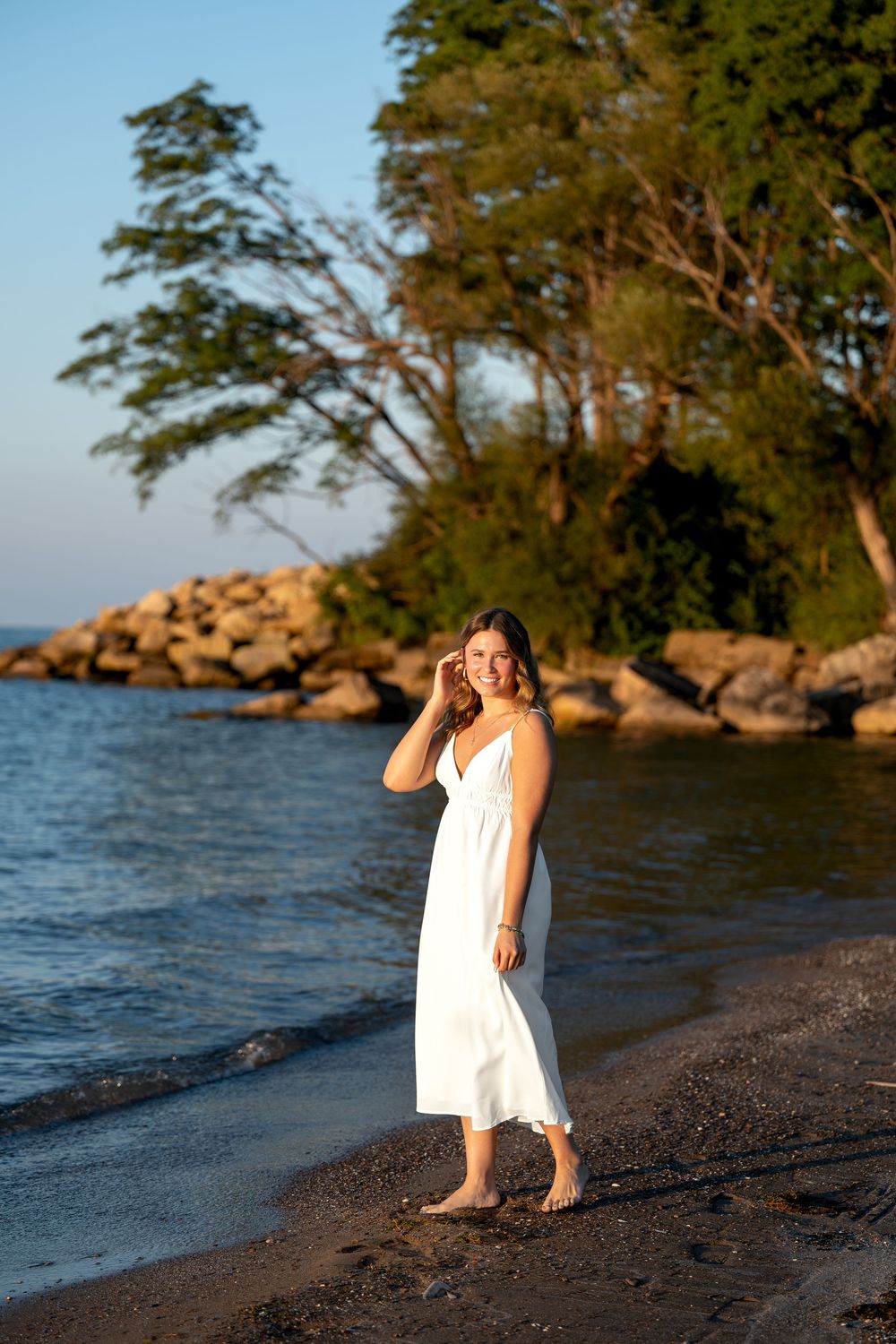 Young woman in white sundress standing barefoot on rocky beach shore at golden sunset hour.