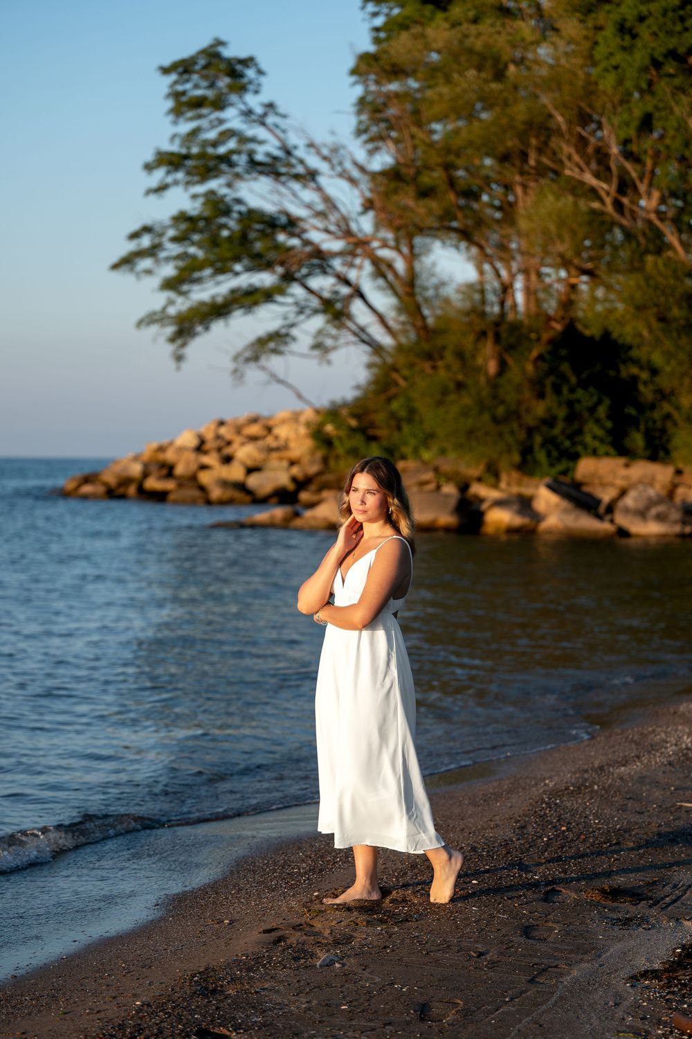 Woman in white dress standing barefoot on rocky beach at golden hour with trees and calm water.