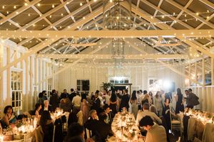 Elegant wedding reception in a greenhouse venue with string lights, candlelit tables, and guests mingling under a white lattice ceiling.
