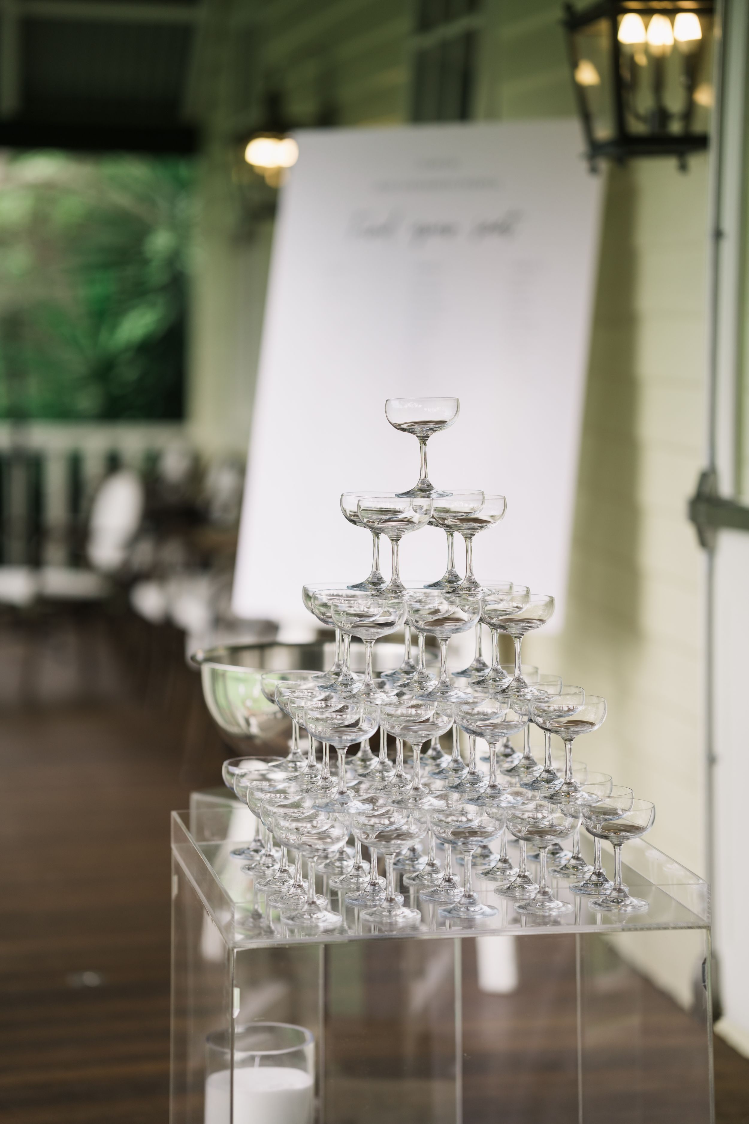 Elegant champagne tower with coupe glasses stacked in pyramid shape on acrylic table at wedding reception.
