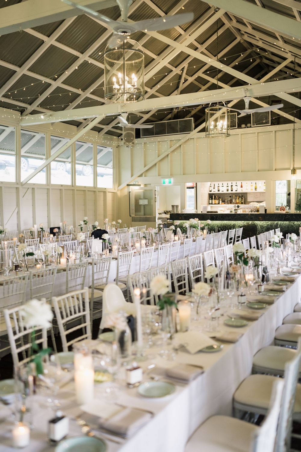 Elegant wedding reception hall with long white tables, silver Chiavari chairs, candles, and floral centerpieces under a vaulted ceiling.