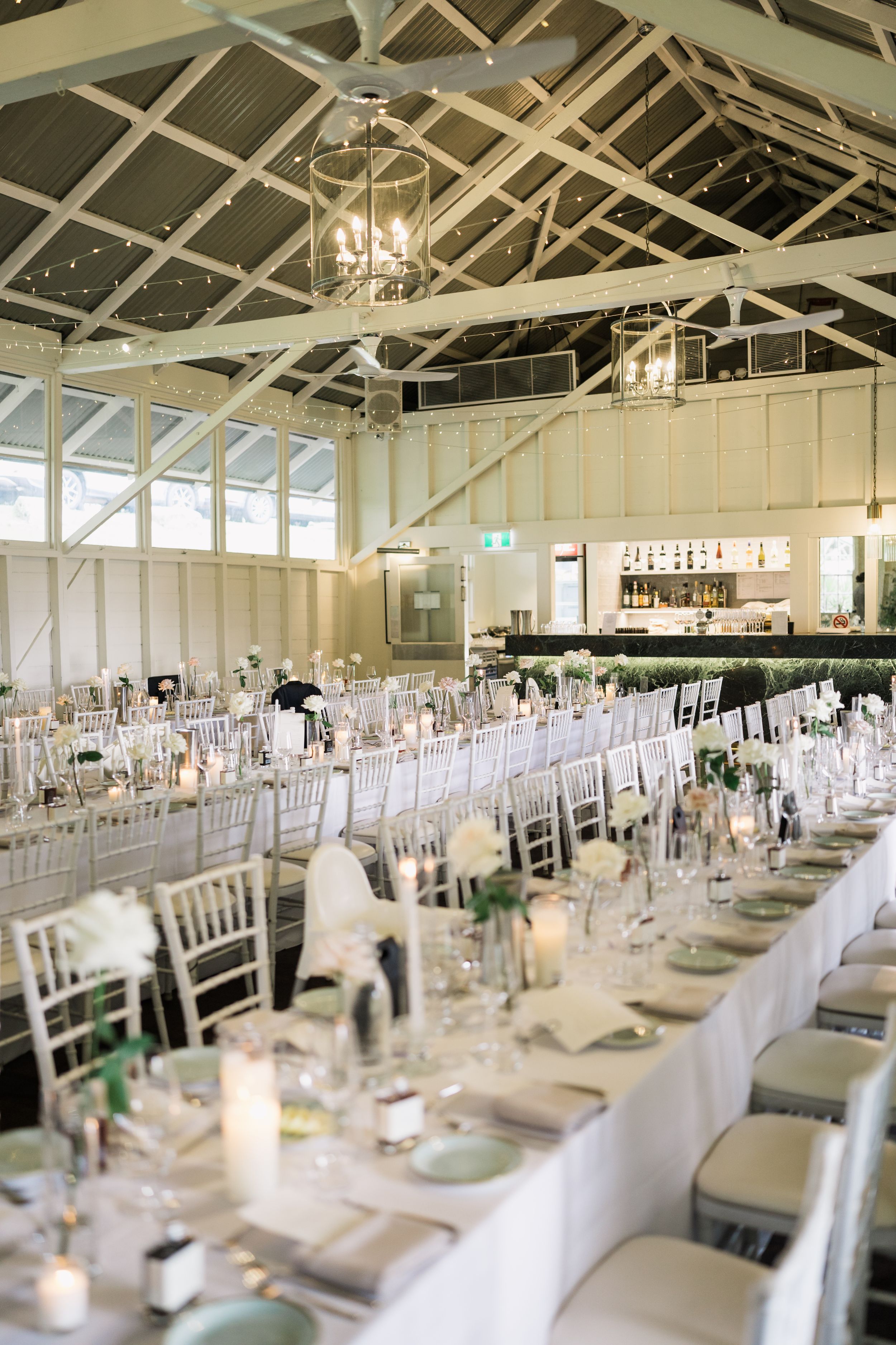 Elegant wedding reception hall with long white tables, silver Chiavari chairs, candles, and floral centerpieces under a vaulted ceiling.