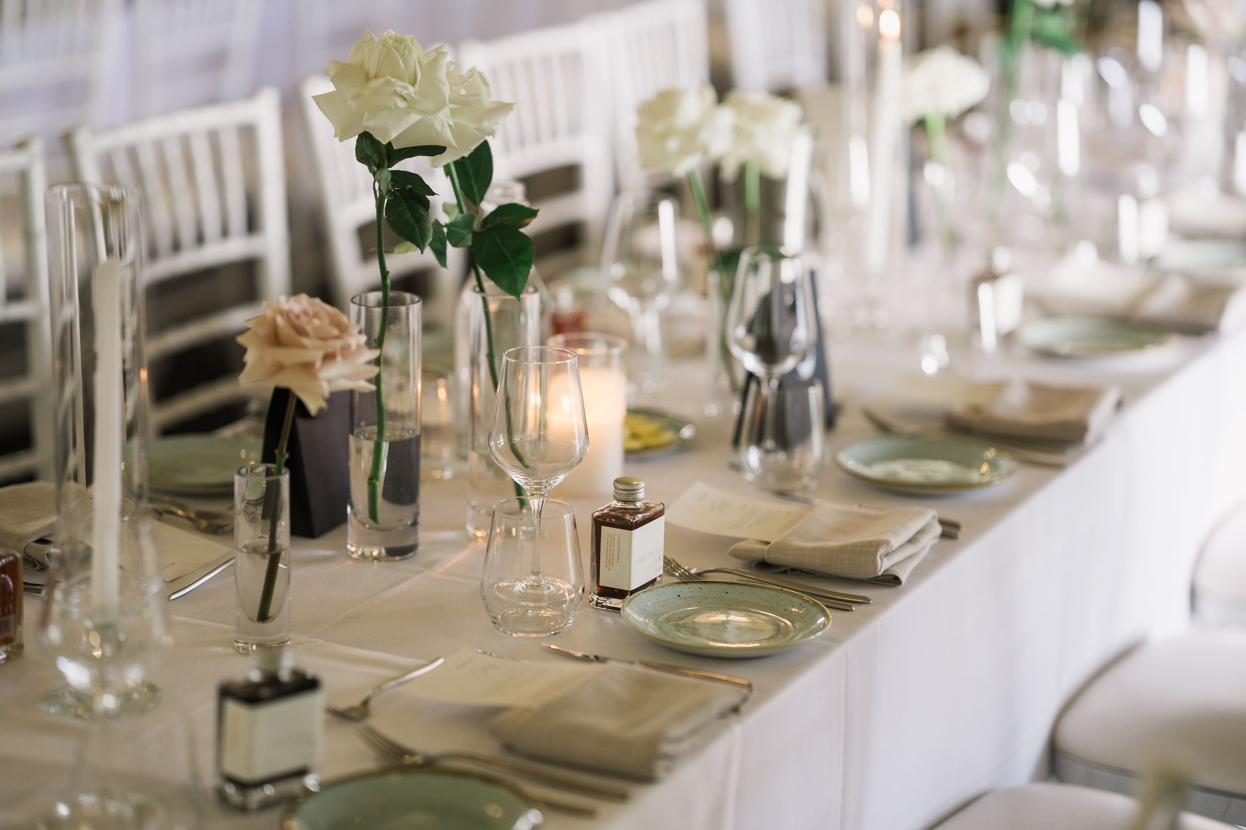 Elegant wedding reception table setting with white roses, candles, and green plates on a white linen tablecloth.
