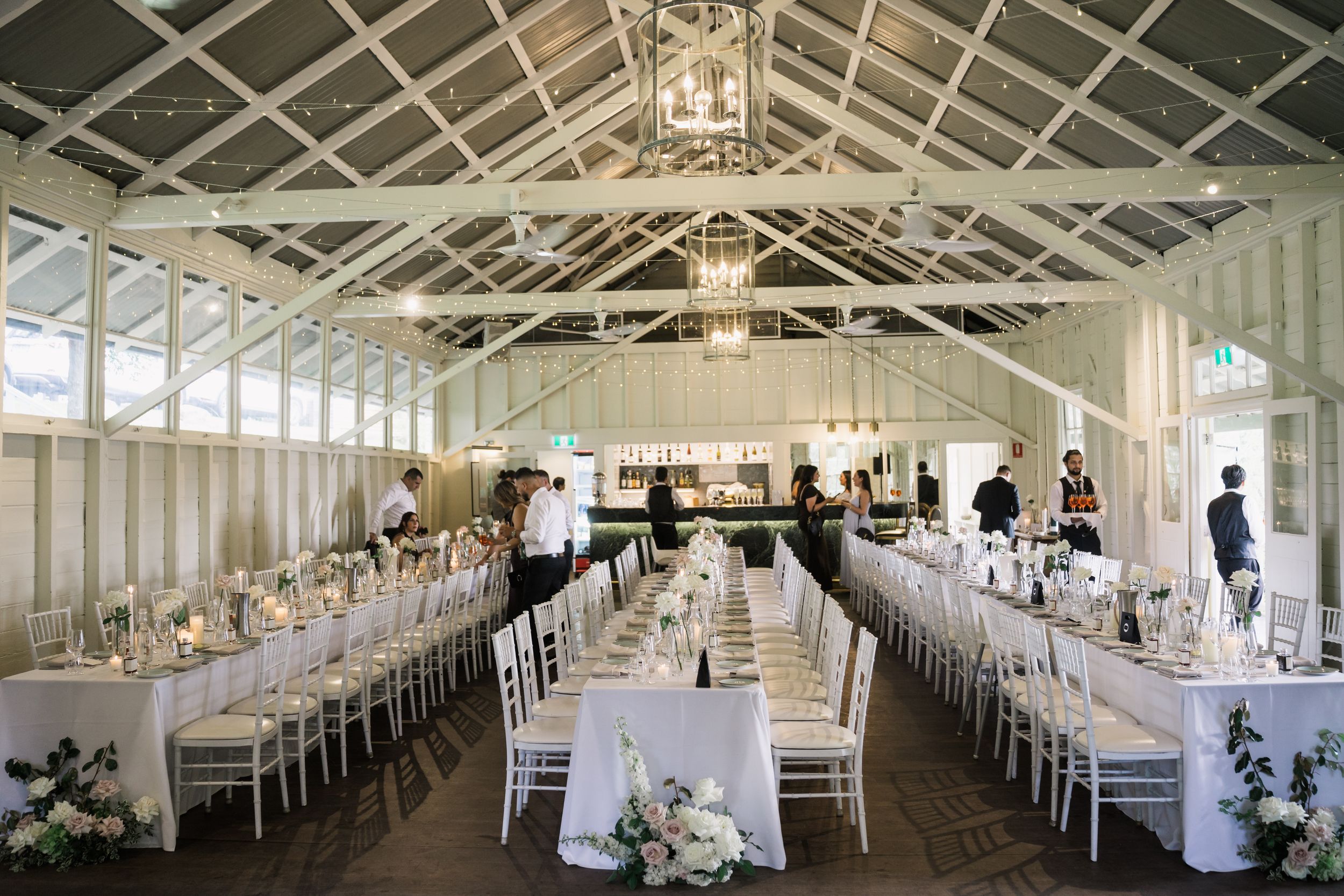 Elegant wedding reception in a bright greenhouse venue with long white-clothed tables, floral centerpieces, and chandelier lighting.