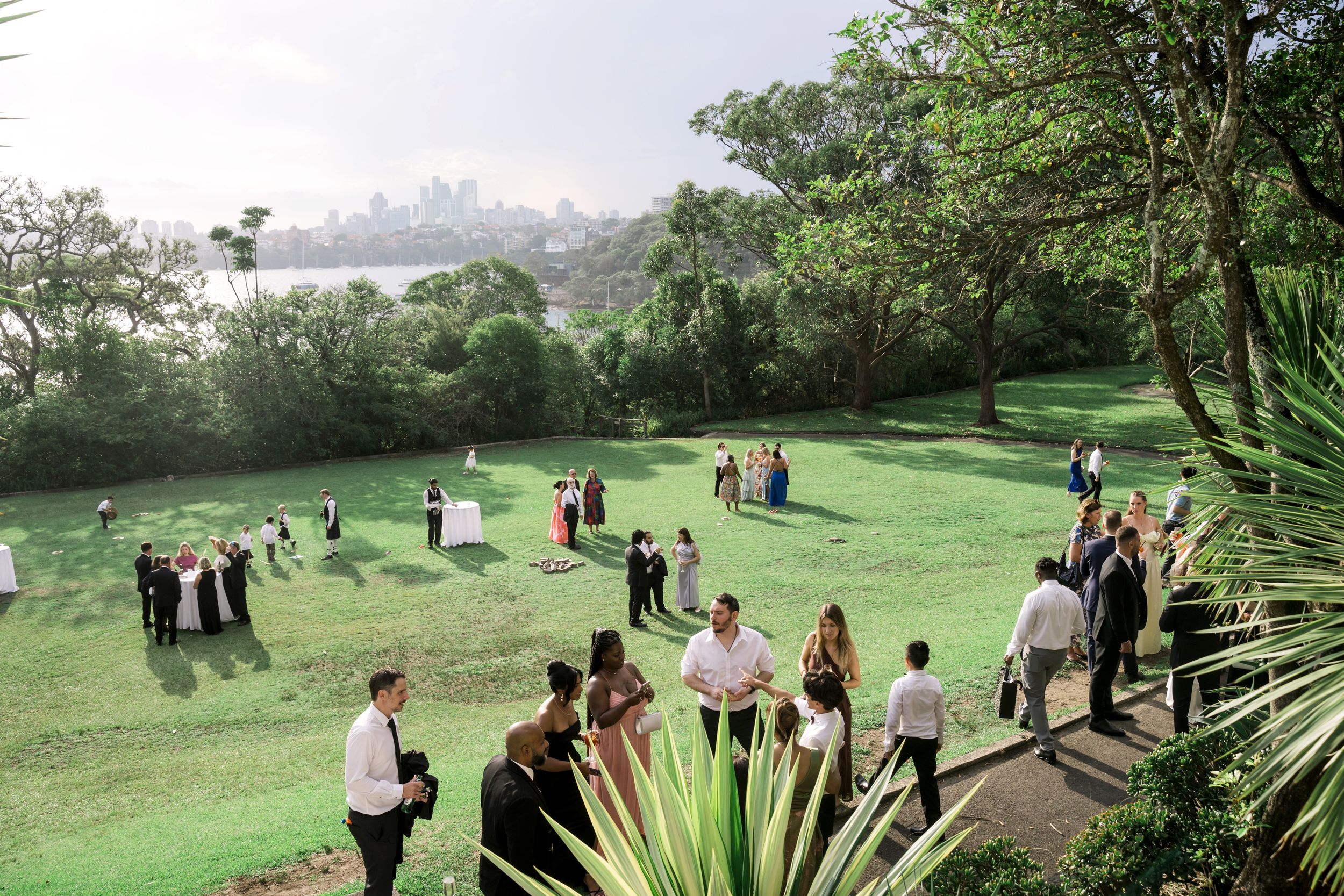 Outdoor wedding reception on lush green lawn with city skyline views, guests mingling among tropical gardens.