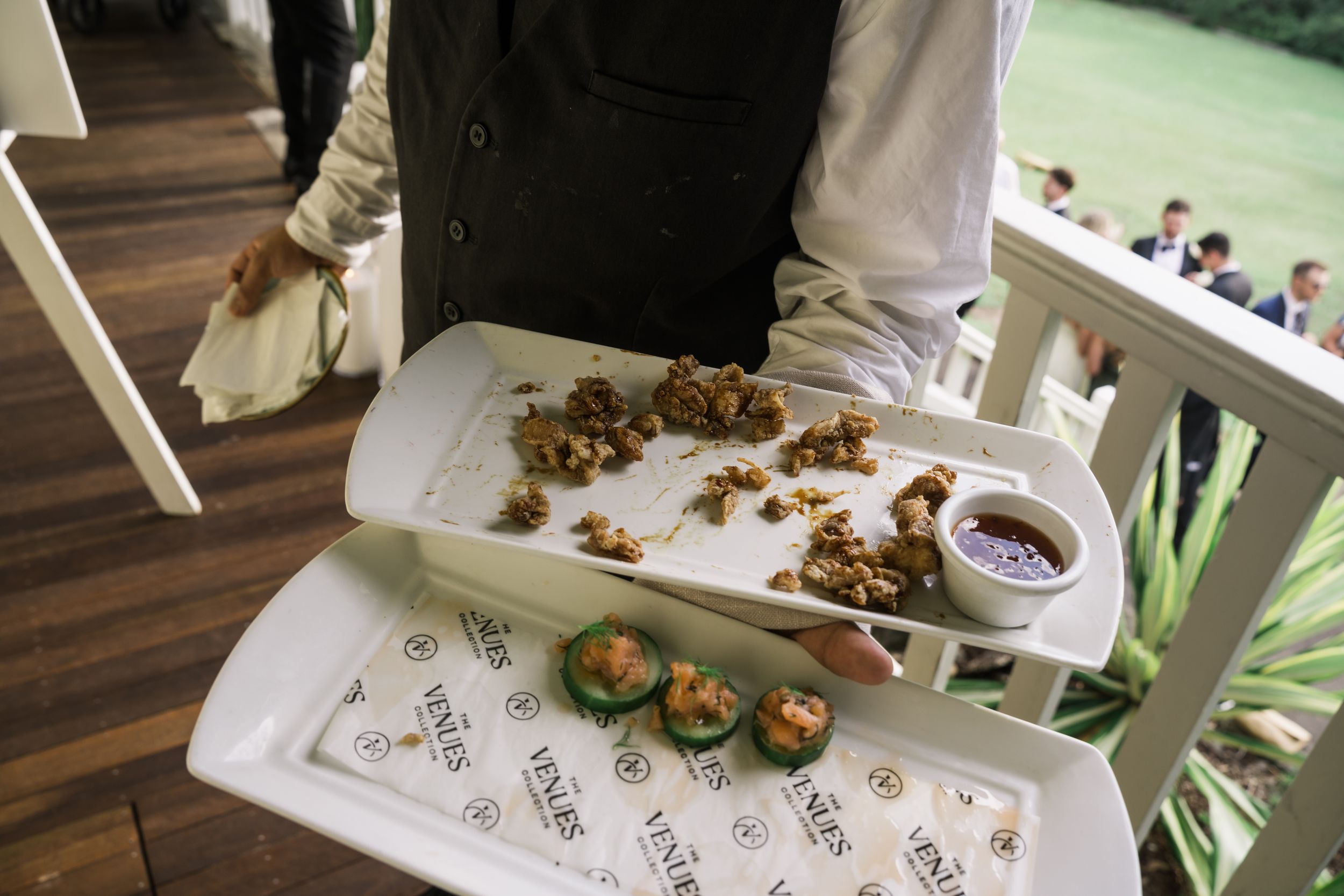 Waiter holding a white tray with gourmet appetizers and dipping sauce at an outdoor catered event.
