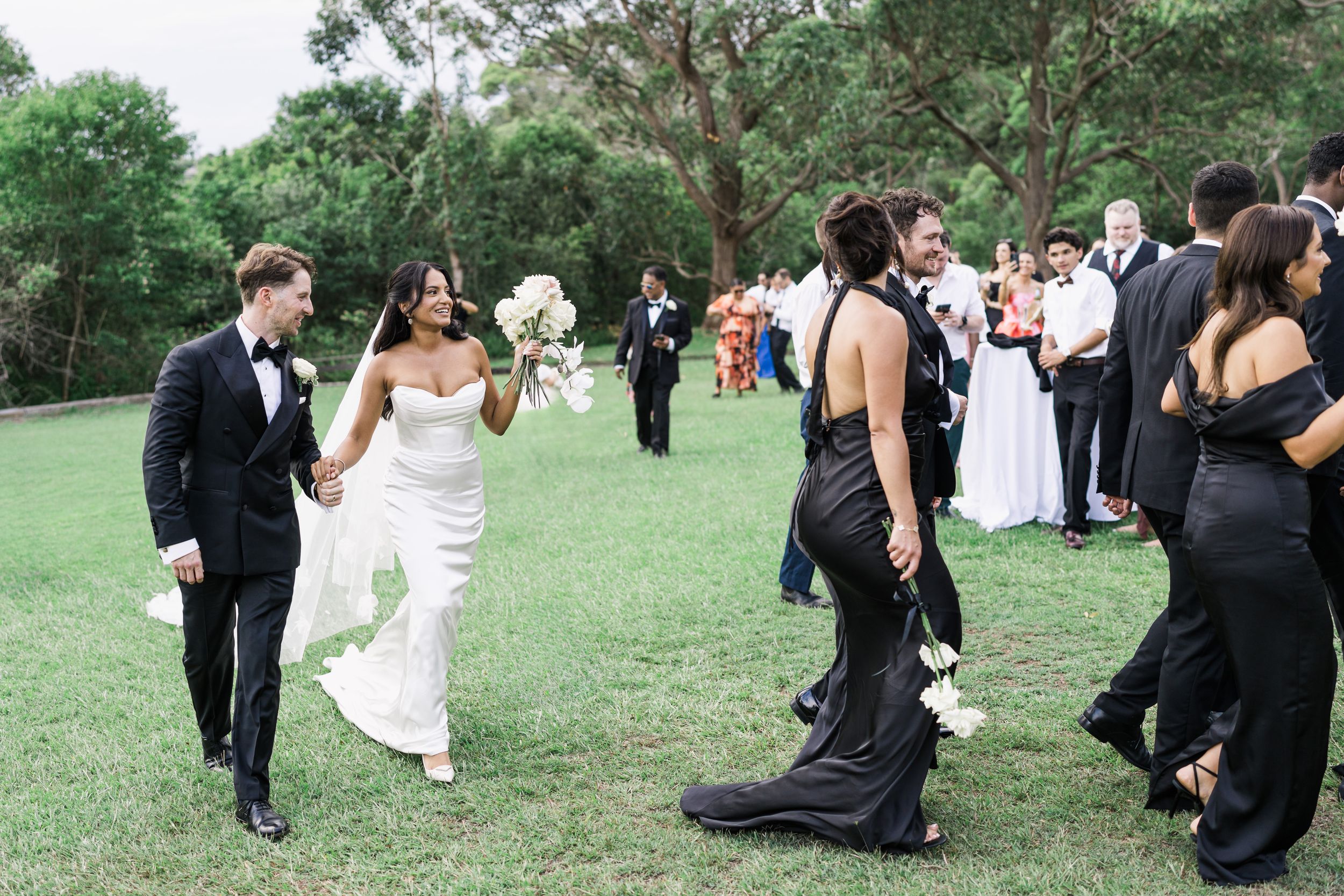 Bride and groom walk hand-in-hand through cheering guests outdoors, bride in white gown holding bouquet, guests in black attire.