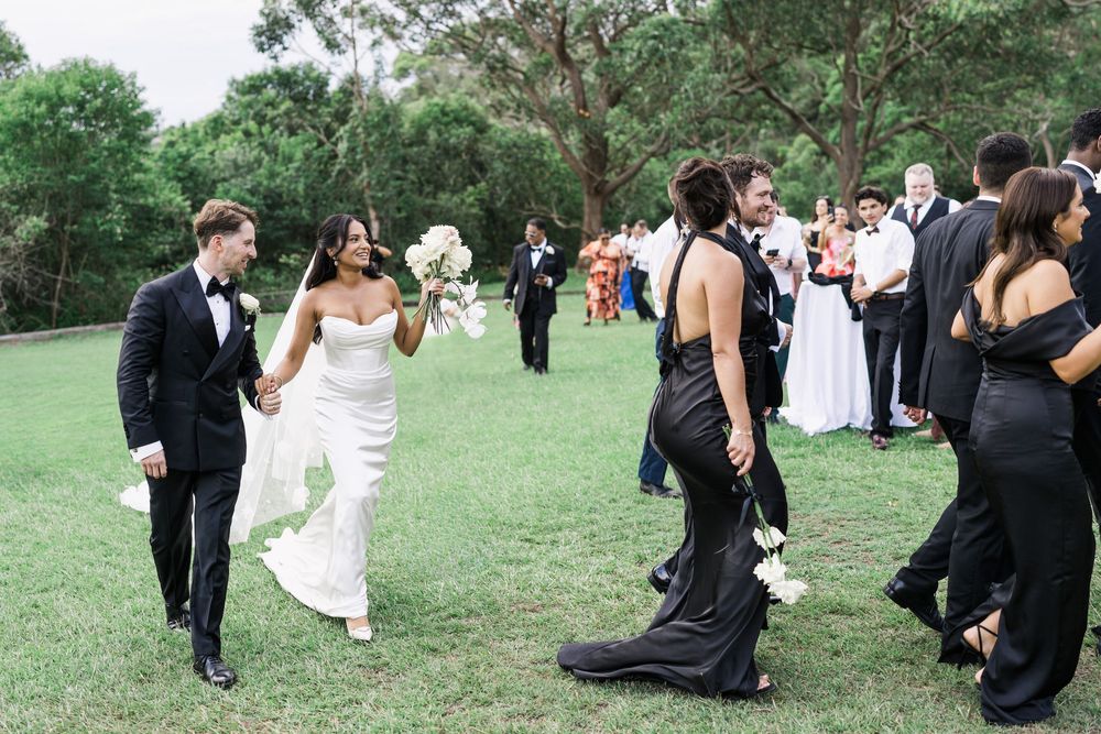 Bride and groom walk hand-in-hand through cheering guests outdoors, bride in white gown holding bouquet, guests in black attire.