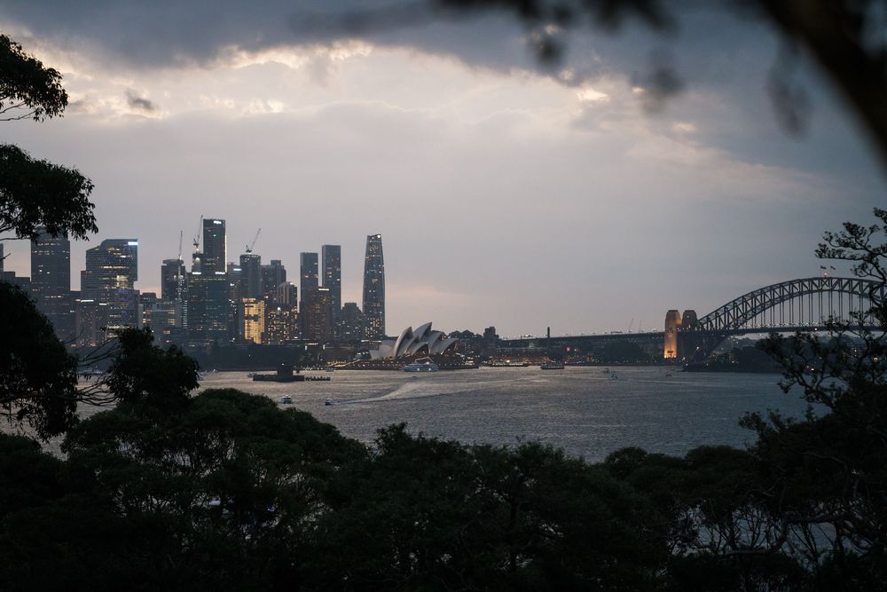 Sydney Harbour skyline at dusk with Opera House, Harbour Bridge, and stormy clouds over calm waters.